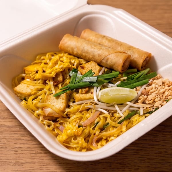 Thai noodle stir-fry with tofu, bean sprouts, chives, lime, crushed peanuts, and spring rolls in a takeout container