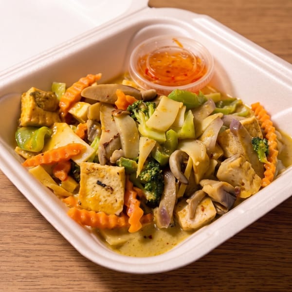 Takeout container filled with tofu, broccoli, carrots, mushrooms, and mixed vegetables in sauce, served with orange dipping sauce.