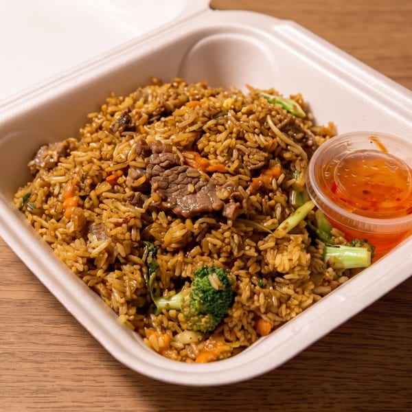 Takeout container filled with beef fried rice, vegetables, and a side of orange dipping sauce on a wooden table.