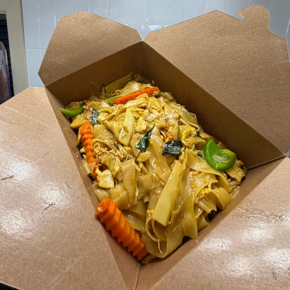 Wide rice noodles stir-fried with chicken, carrots, peppers, and leafy greens in an open cardboard takeaway box.
