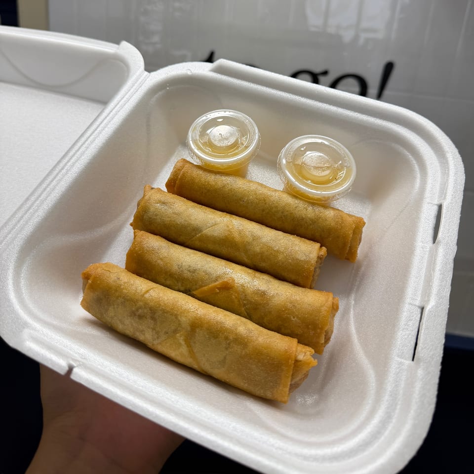Golden fried spring rolls arranged in a foam takeout container with two small cups of dipping sauce.