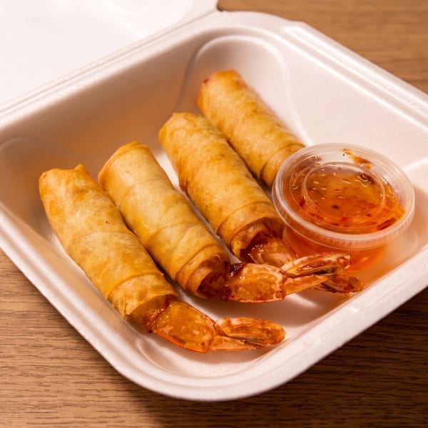 Four golden fried spring rolls with shrimp tails and sweet chili dipping sauce in a takeout container.